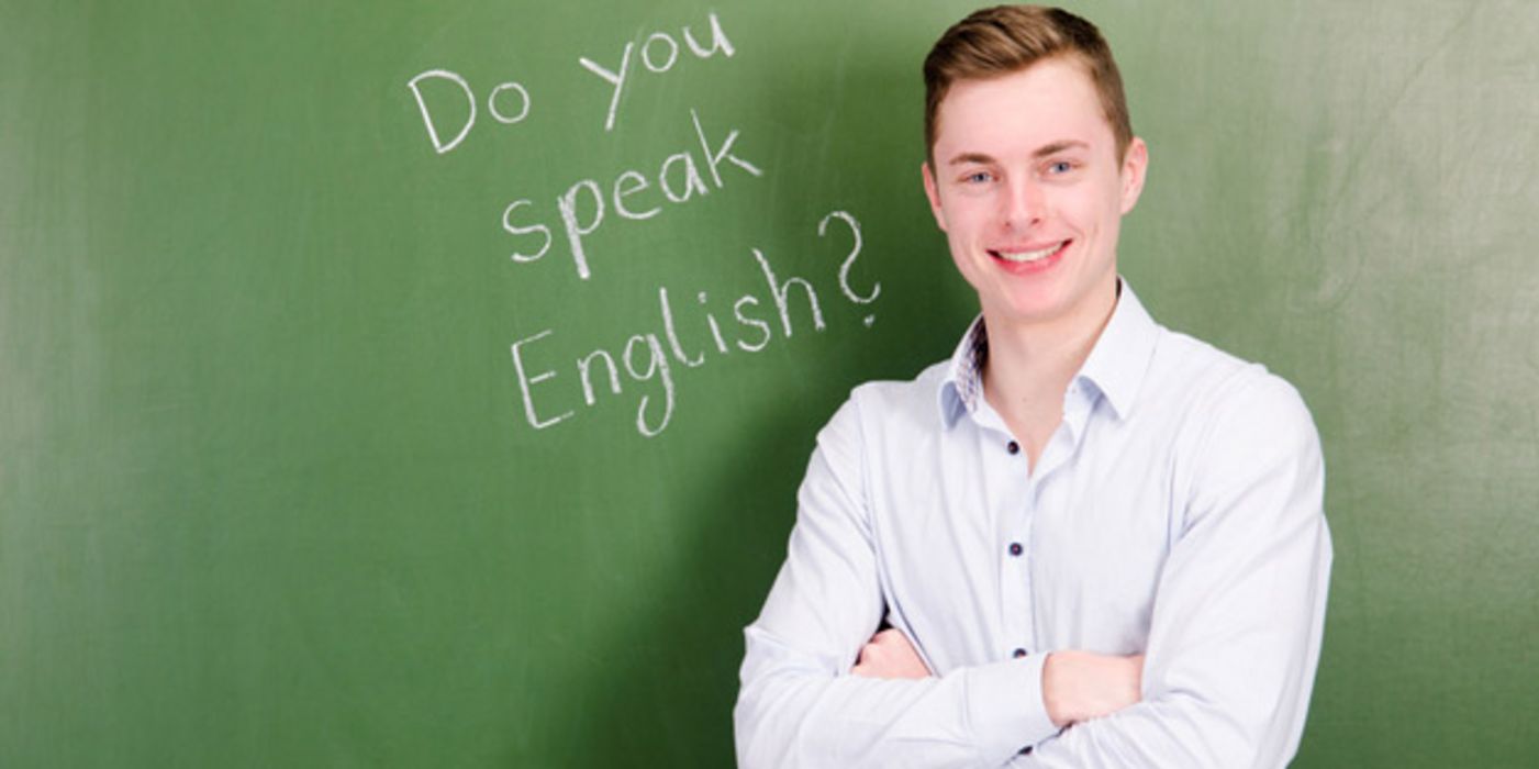 Gymnasiast, ca. 17, 18 Jahre, helles Hemd, lachend, verschränkte Arme, mit dem Rücken zu eine Schultafel, auf der zu lesen ist "Do you speak English?"