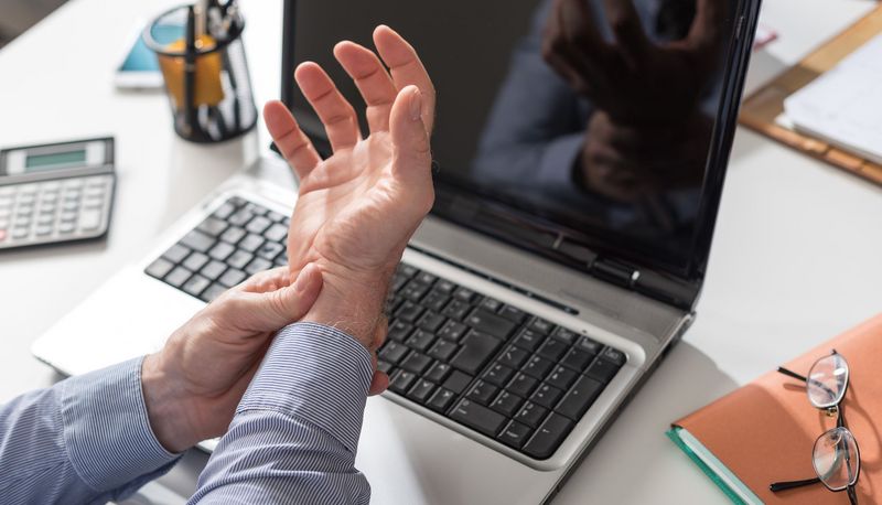 Mann hält sein Handgelenk, im Hintergrund ein Laptop.