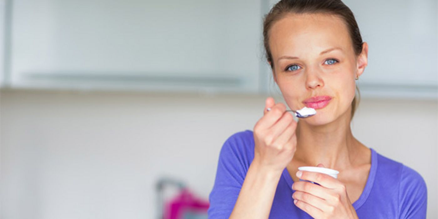 Frontalfoto Frau ca. 30 in lilablauem Shirt, blaue Augen, rosa geschminkte Lippen, Haare zurückgebunden, löffelt genussvoll einen Joghurt