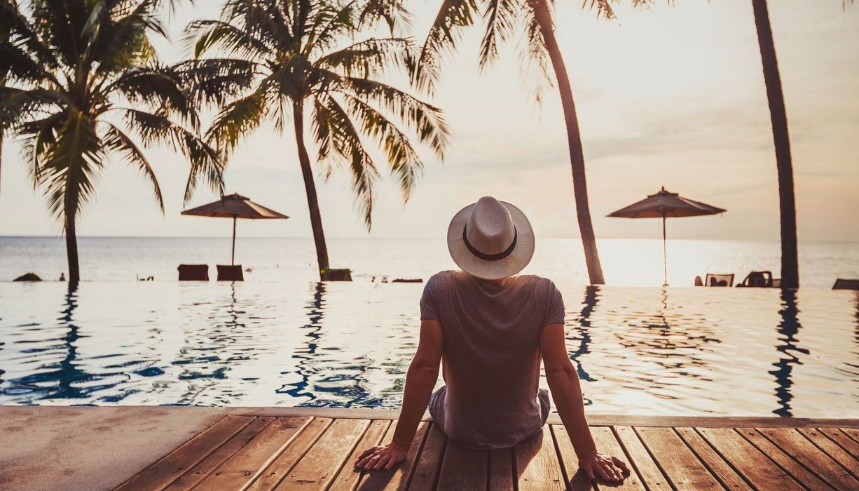 Mann mit Hut, sitzt vor einem Pool mit Blick auf das Meer.