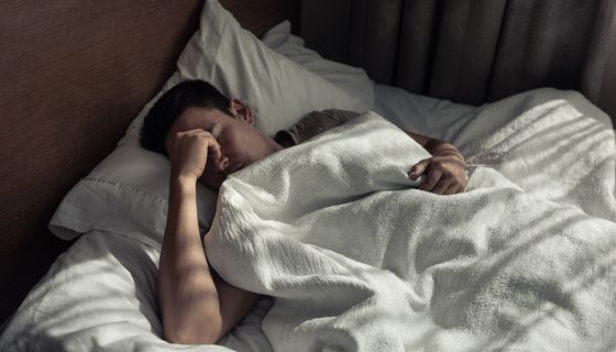 Mann im Bett am morgen: Er wirkt unausgeruht und greift sich mit einer Hand an den Nasenrücken.