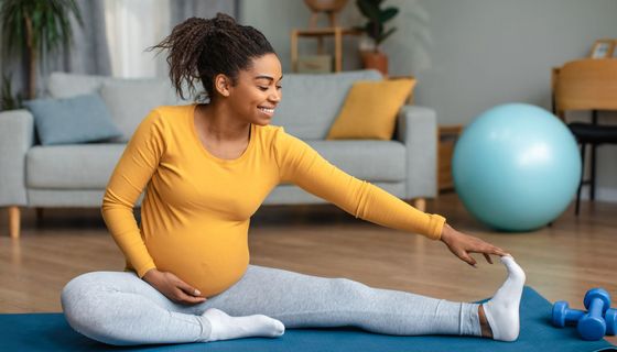 Schwangere Frau macht Dehnübungen im Sitzen auf einer Matte. Am Fußende liegen Hanteln.