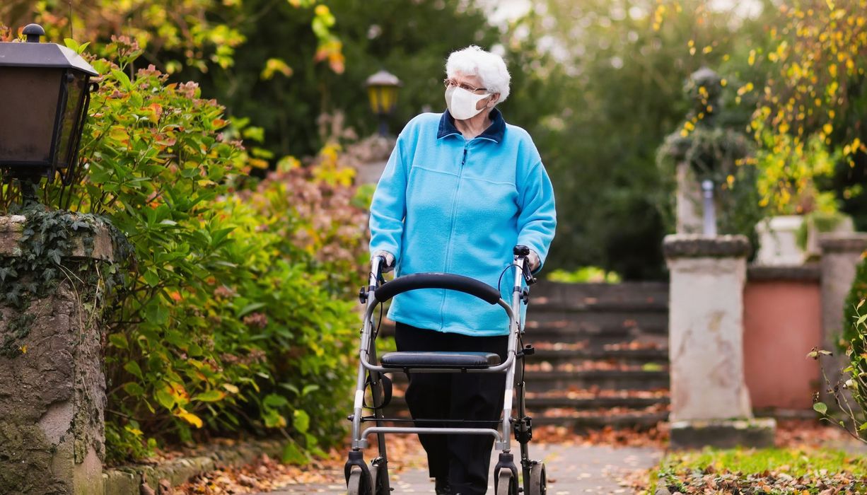 Ältere Frau mit Maske, geht draußen am Rollator spazieren.