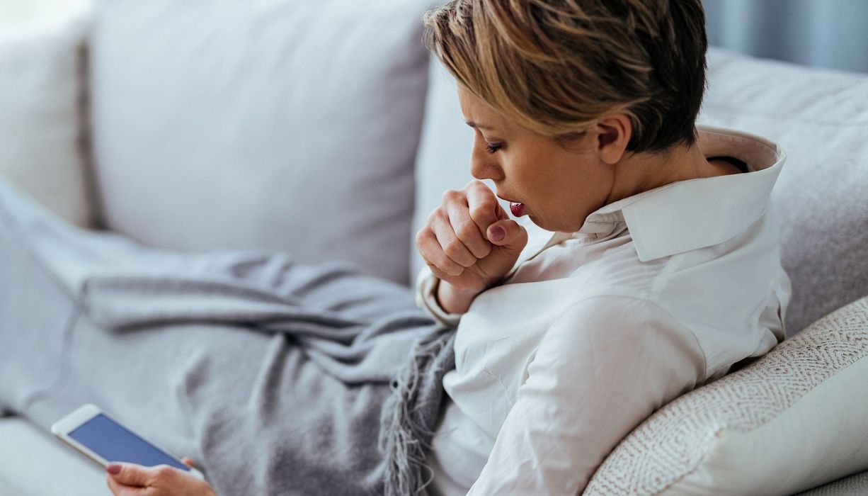 Junge Frau, liegt auf der Couch und hustet, mit Smartphone in der Hand.