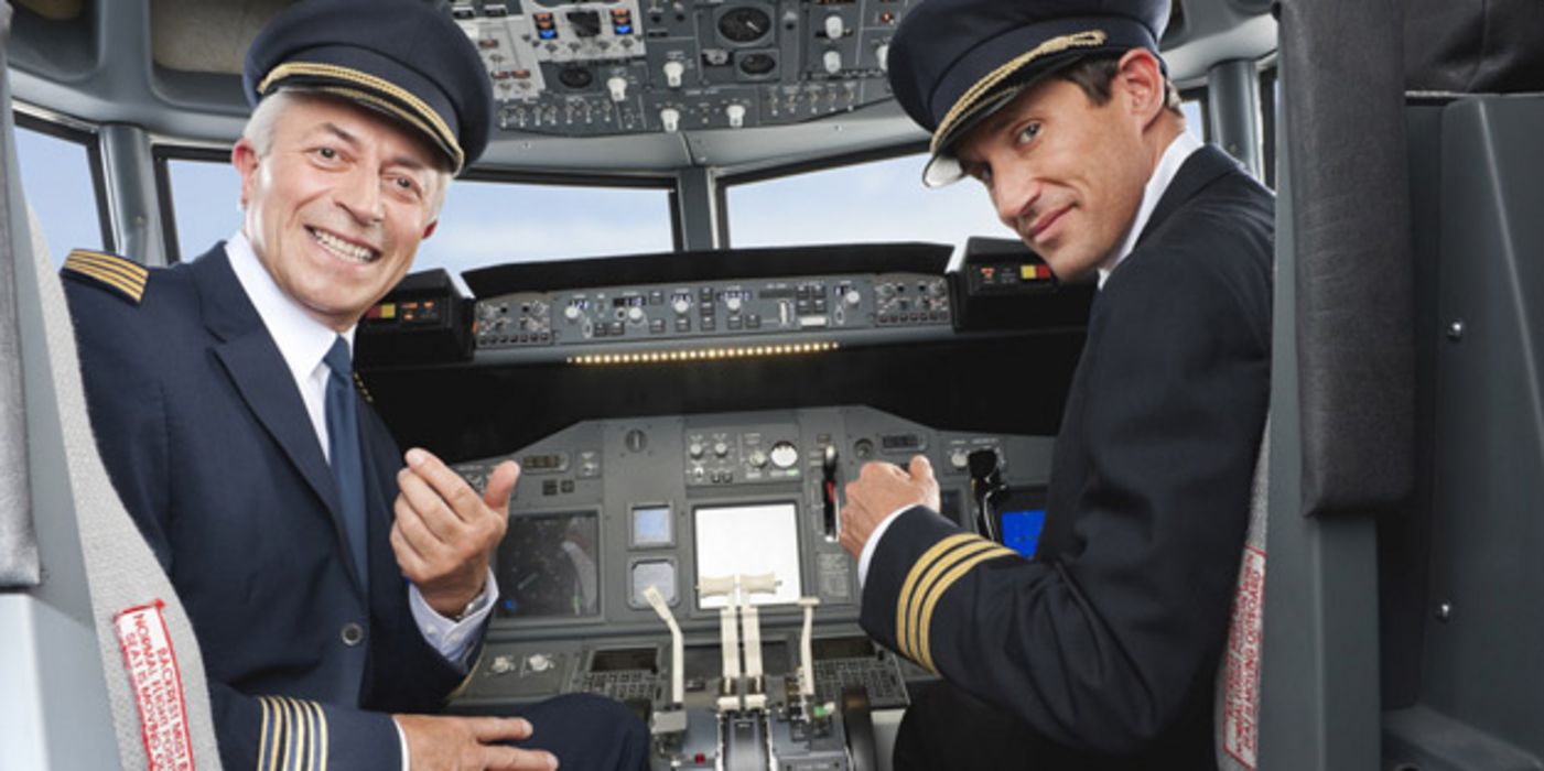 Zwei Piloten im Cockpit schauen aus ihren Sitzen heraus über die Schulter in die Kamera und lächeln