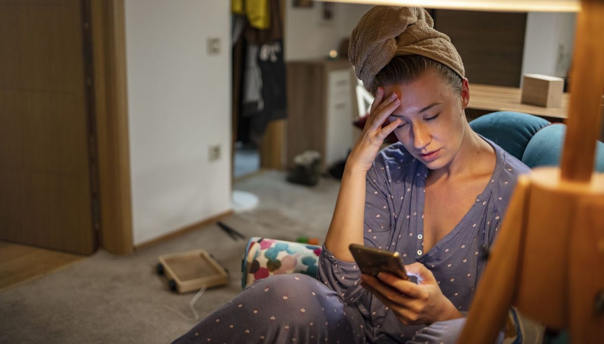 Junge Frau, zu Hause und krank, hält ihr Handy in der Hand.