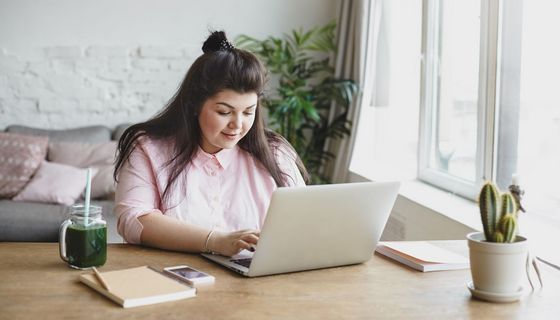Eine übergewichteige junge Frau sitzt vor einem Laptop und tippt.