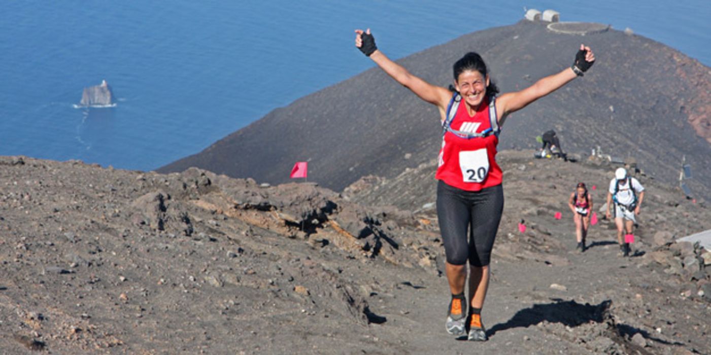 Ultra-Marathon-Läuferin auf Berggipfel, im Hintergrund das Meer, reißt freudig die Arme hoch