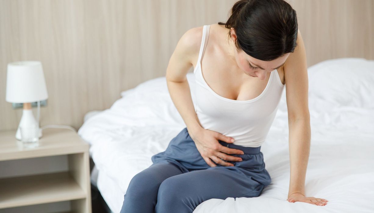 Junge Frau sitzt auf dem Bett und hält sich den Unterleib.