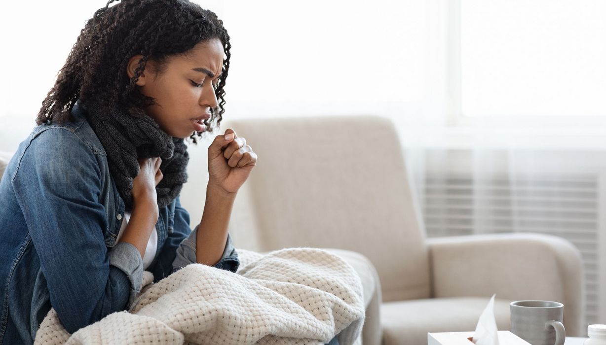 Junge Frau, sitzt auf der Couch und hustet.