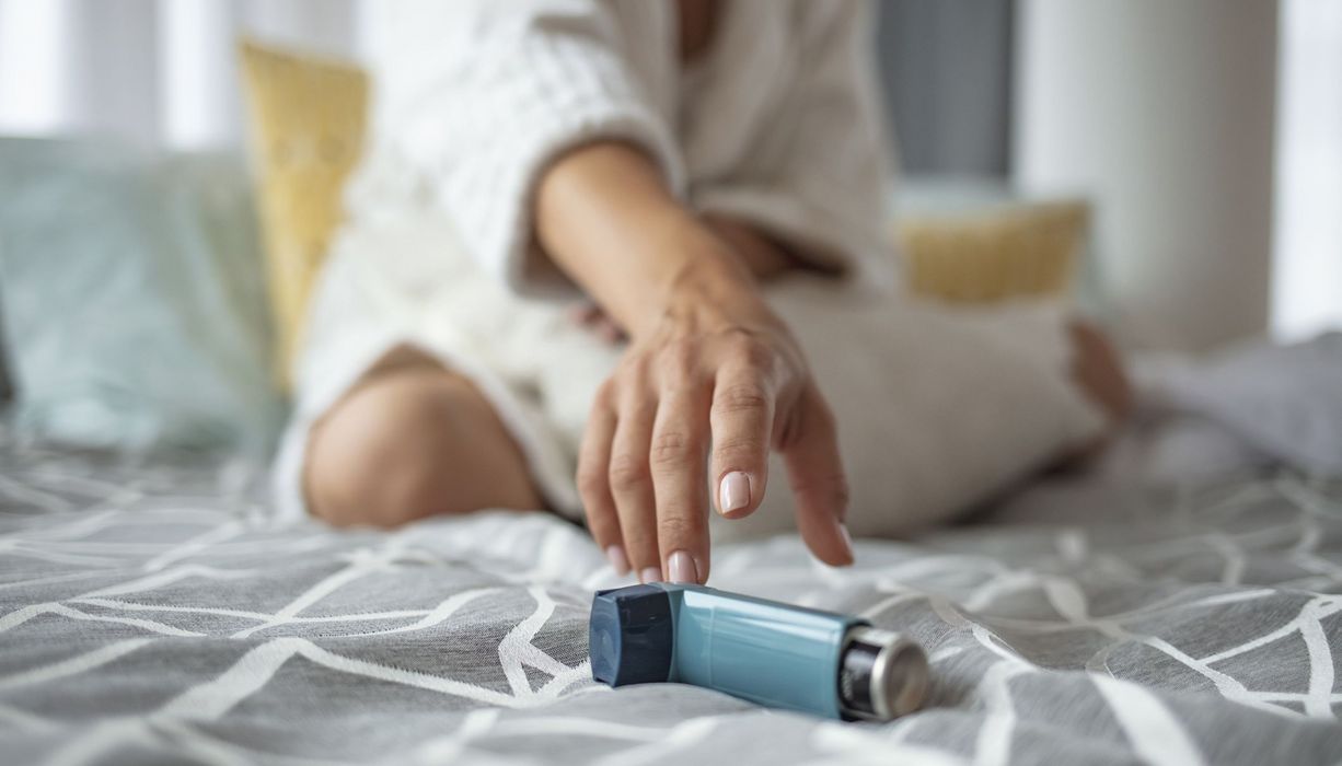 Junge Frau, sitzt im Bett und greift nach Asthmaspray.