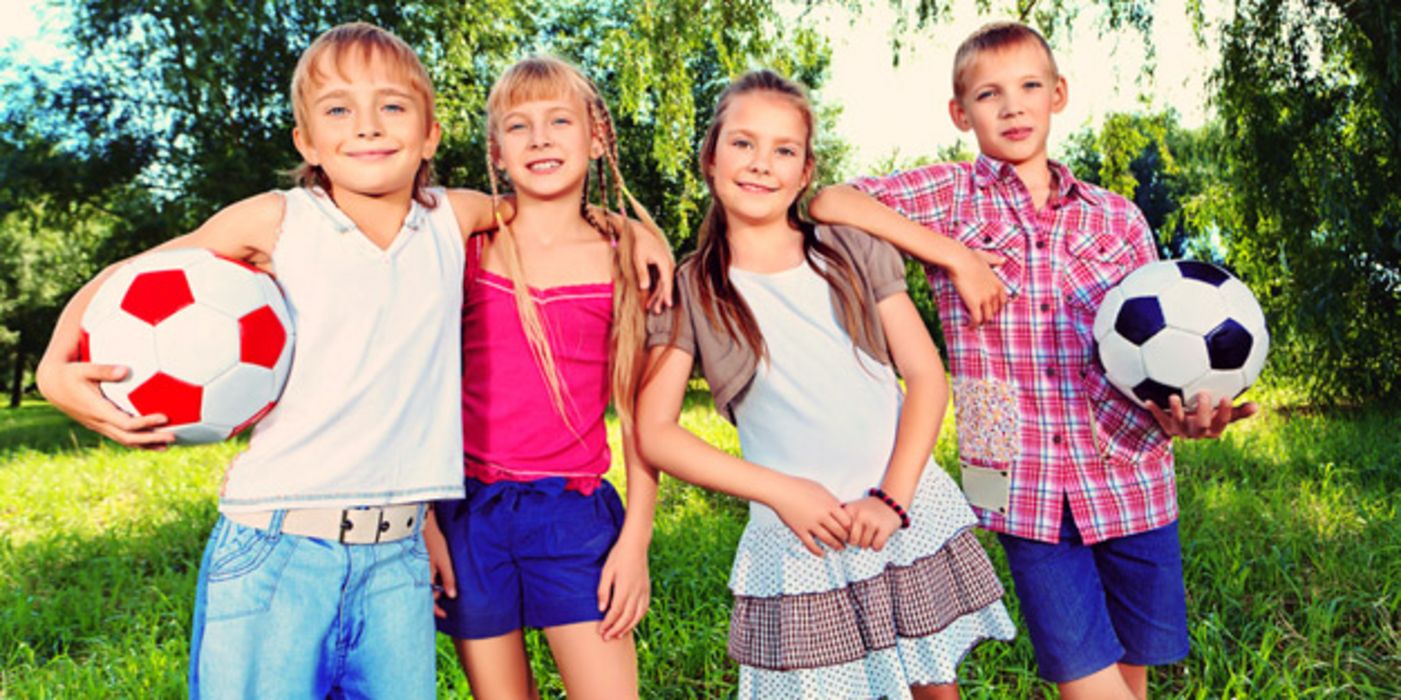 Zwei Jungen und zwei Mädchen stehen in sommerlichen Outfits im Freien, die Jungen tragen je einen Fußball unter dem Arm