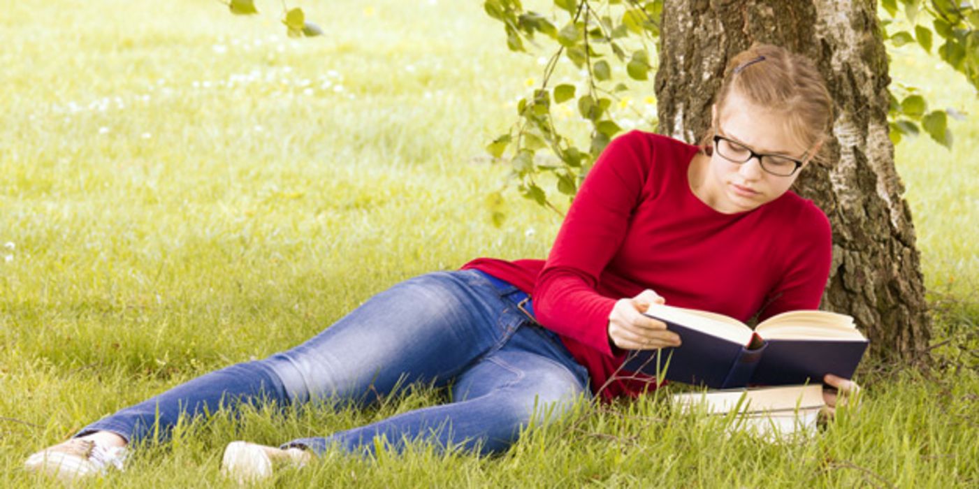 Junge Frau liegt unter einer Birke im Gras und liest ein Buch.