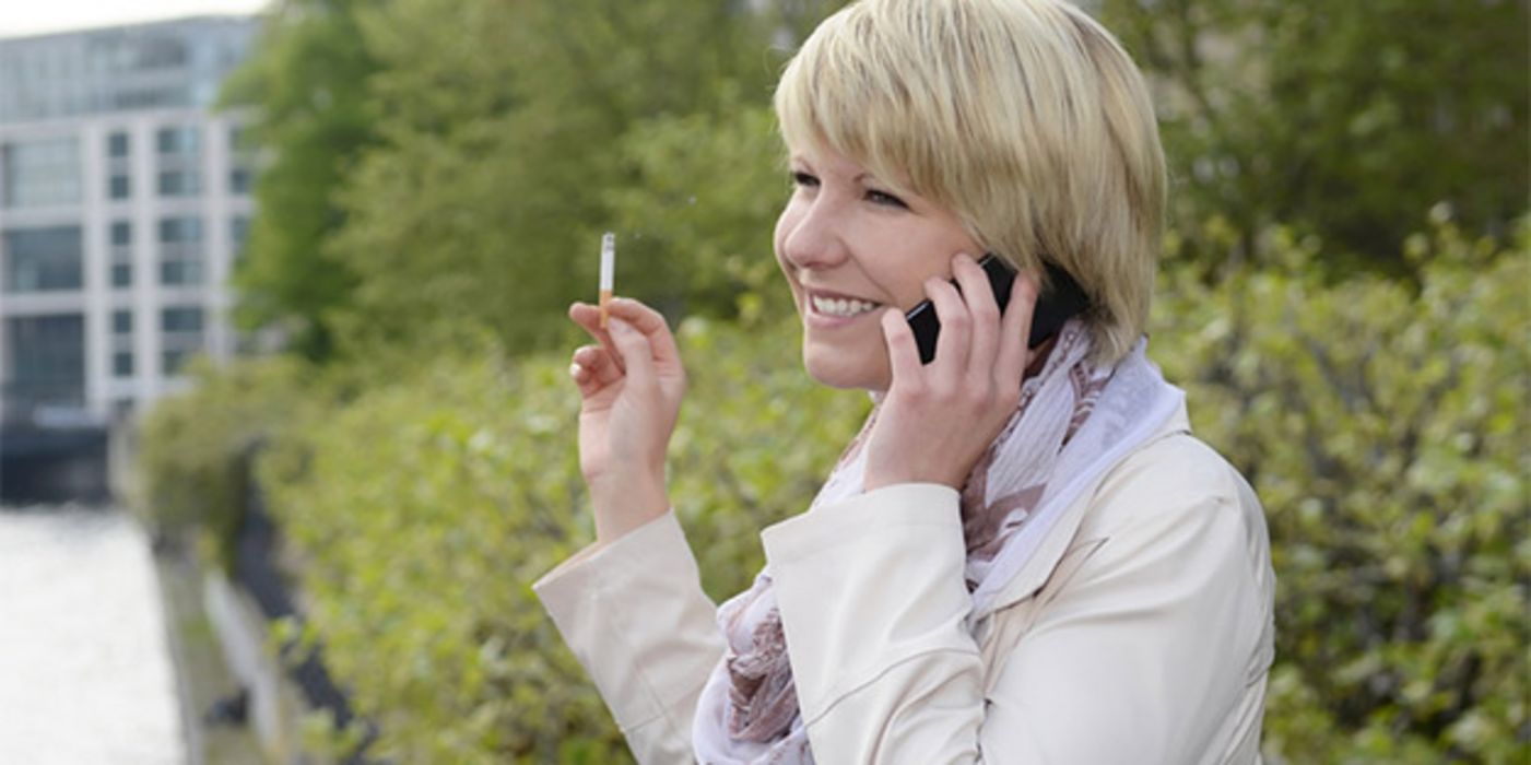 Frau in den 40ern, Profilbild, blond, Zigarette, Handy, vor Busch, Hintergrund Häuser, heller Trenchcoat