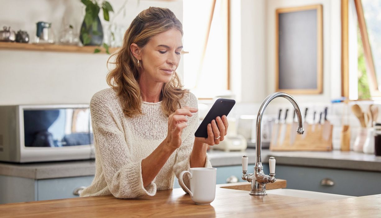 Frau steht in der Küche und schaut auf ihr Smartphone.