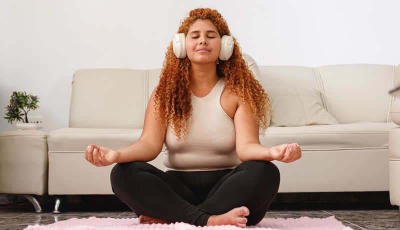 Frau sitzt in Zimmer auf einem Teppich vor einer Couch. Sie trägt Kophörer und hat geschlossene Augen.