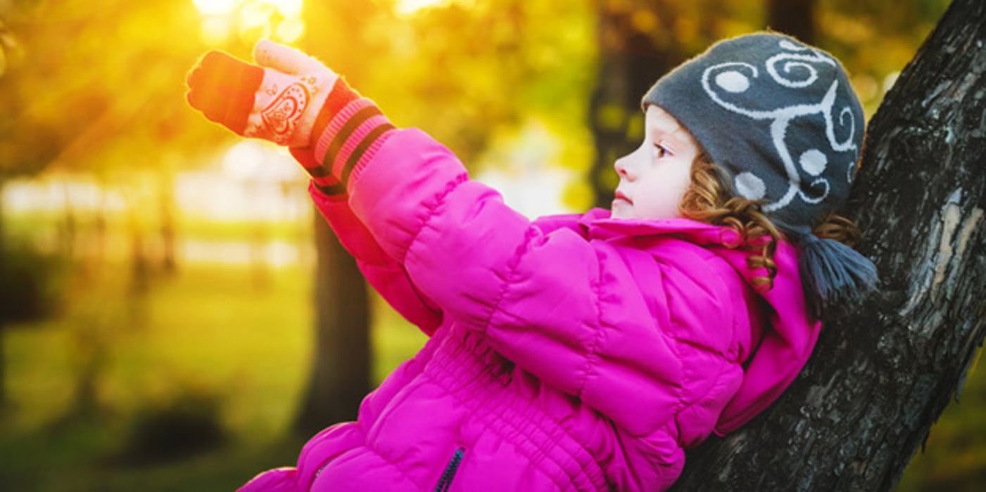 Mädchen, ca. 7 Jahre, zyklamfarbener Anorak, blauweiße Mütze, im Herbstabendlicht an einen Baum gelehnt