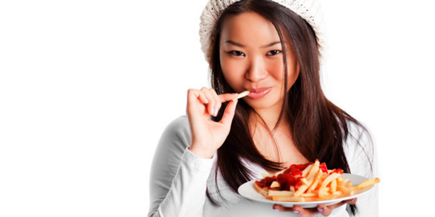 Junge Asiatin mit Strickmütze hält einen Teller mit Pommes frites in der Hand und nascht einen davon