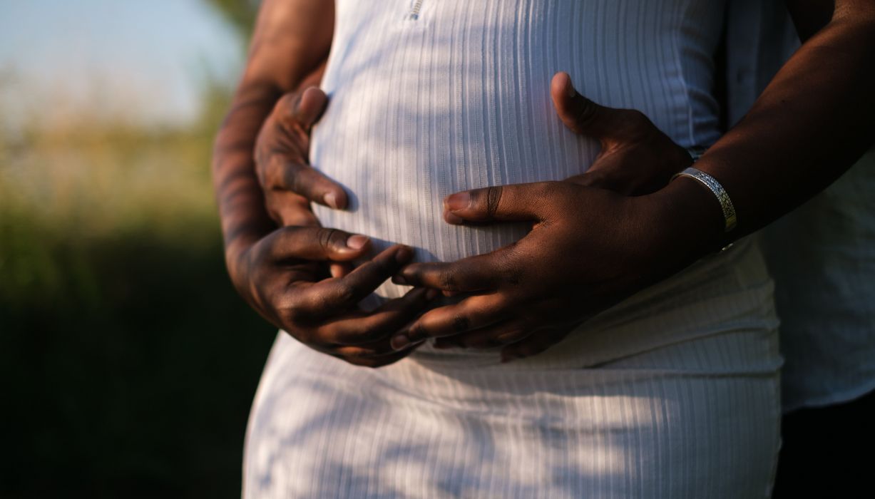 Frau mit Babybauch, wird von hinten von einem Mann umarmt, der seine Hände auf den Bauch legt.