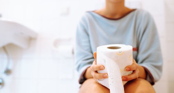 Frau, sitzt auf einer Toilette und hält eine Rolle Toilettenpapier in der Hand.