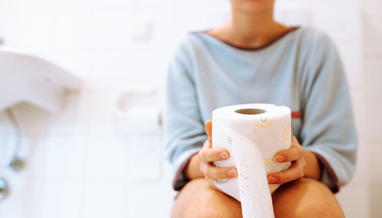 Frau, sitzt auf einer Toilette und hält eine Rolle Toilettenpapier in der Hand.