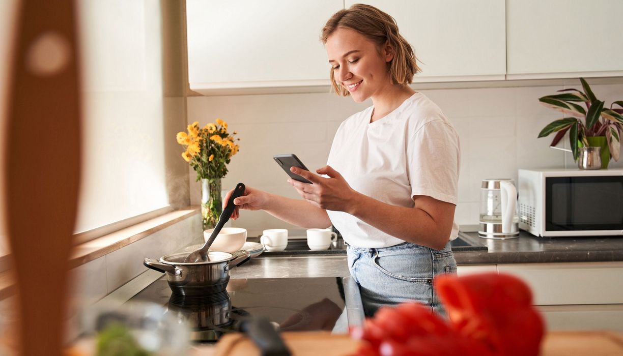 Junge Frau, kocht eine Suppe und schaut auf ihr Smartphone.