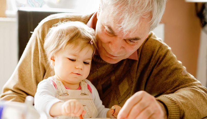 Etwa einjähriges Kind malt ein Bild, gehalten von einem Mann, etwa über 70 Jahre.