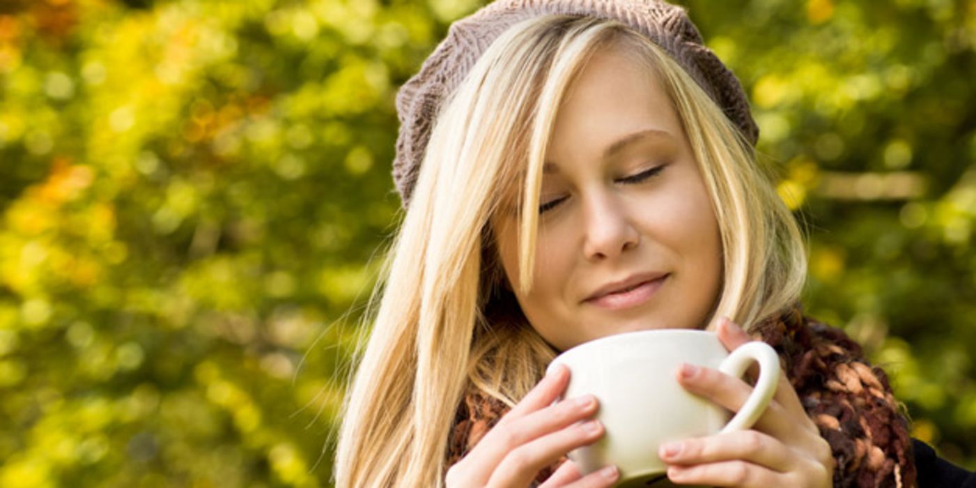 Blonde junge Frau, ca. 18, hellbraune Wollmütze, blonde halblange Haare, schaut genüsslich in eine Kaffeetasse; im Hintergrund herbstlicher Blätterwald