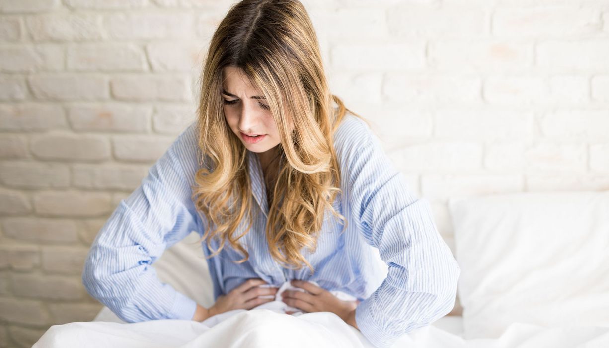 Junge Frau, sitzt im Pyjama in ihrem Bett und hält sich den Bauch.