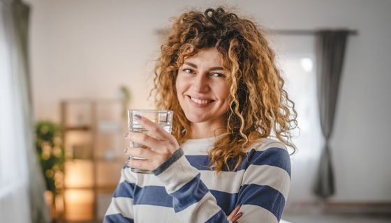 Frau mit blonden Locken hält ein Glas Wasser in der Hand.