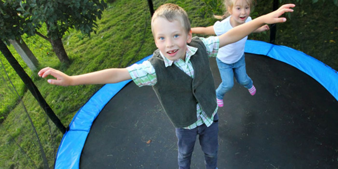 Junge im Vordergrund, Mädchen im Hintergrund hüpfen auf Trampolin und schauen in die Kamera