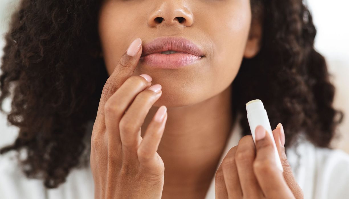 Dunkelhäutige Frau, hält Lippenpflegestift in der einen Hand und trägt diesen mit der anderen Hand auf.