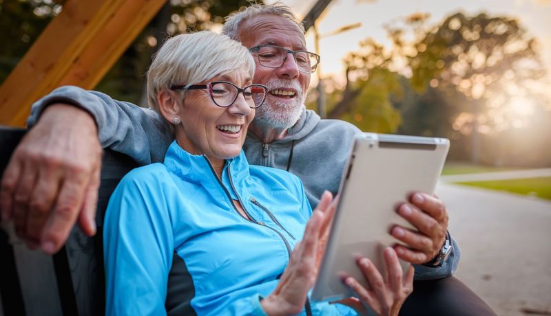 Ein älteres Pärchen, sitzt auf einer Bank und schaut lächelnd auf ein Tablet.