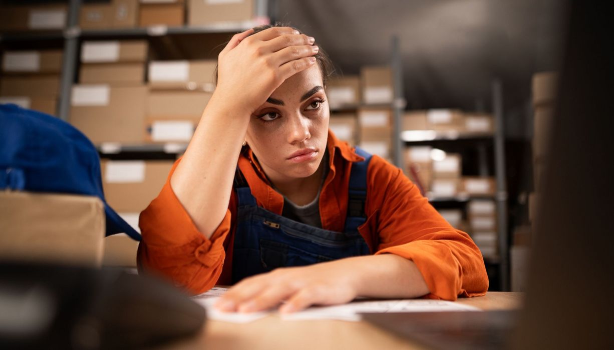 Junge Frau, sitzt bei der Arbeit in einem Lager und stützt die den Kopf in die Hände.