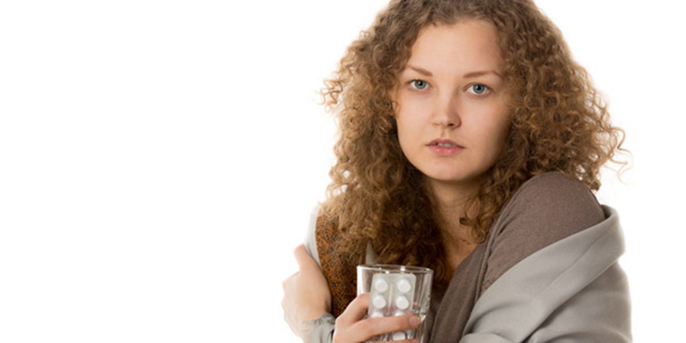 Junge Frau, dunkele, längere Locken, braune Decke um die Schultern, Wasserglas und Blisterpackung in der Hand