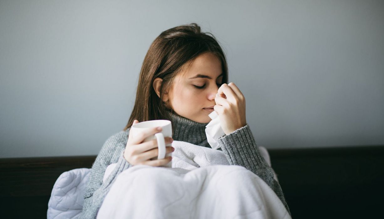 Frau sitzt mit einer Tasse Tee im Bett, hat Schnupfen. 