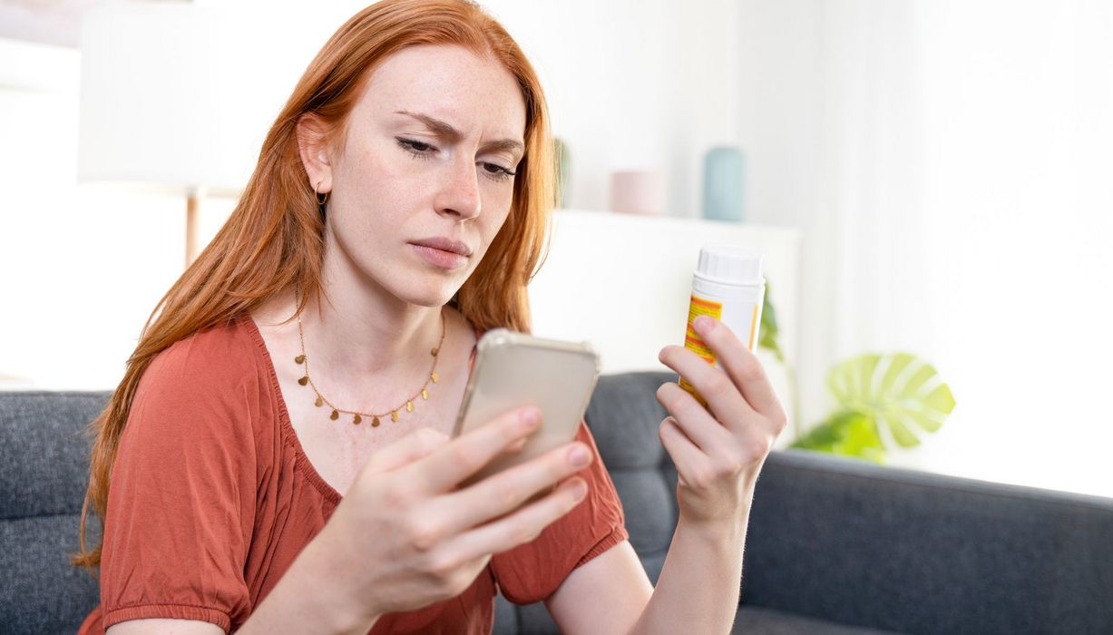 Junge Frau, hält ein Medikament in der Hand und schaut skeptisch auf ihr Handy.