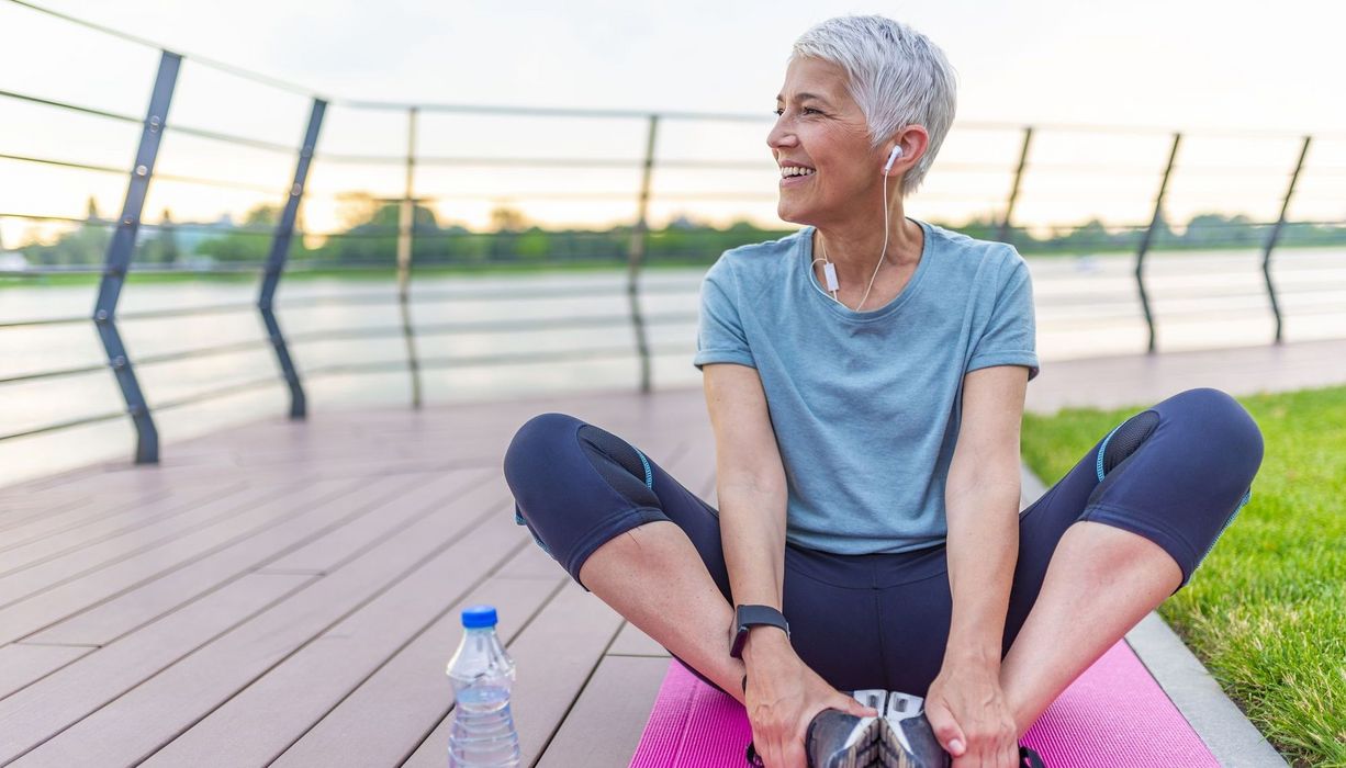 Seniorin in Sportkleidung, sitzt draußen und dehnt sich.