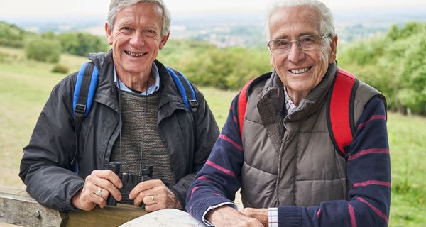 Zwei ältere Männer auf Wandertour lehnen an einem Zaun.