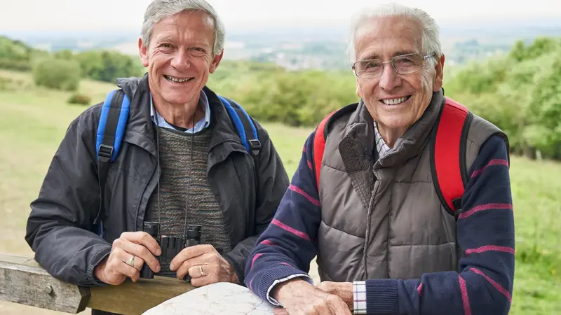 Zwei ältere Männer auf Wandertour lehnen an einem Zaun.