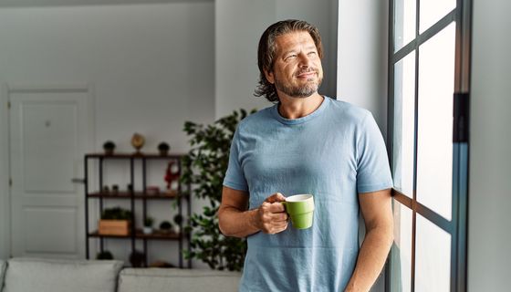 Mann, steht mit einer Kaffeetasse am Fenster.