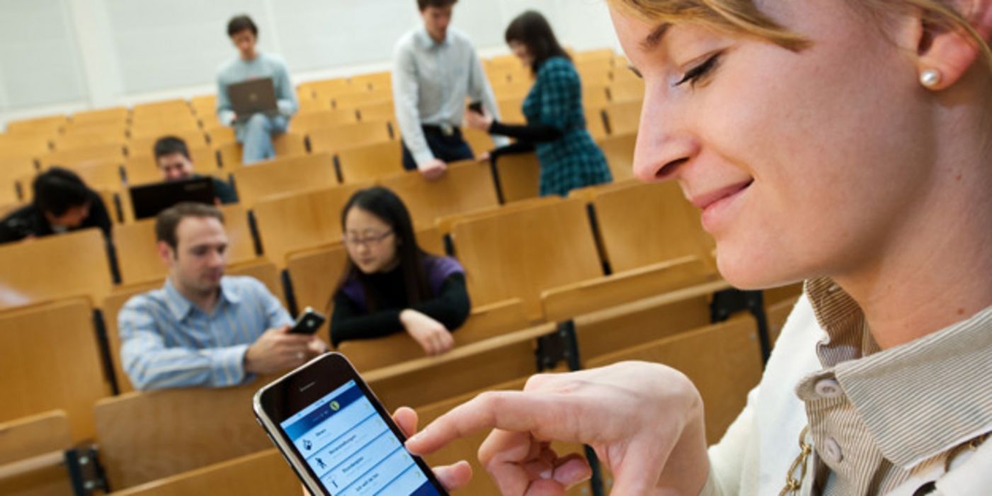 Hörsaal im Hintergrund mit Studierenden, Profil einer Studentin im Vordergrund die auf ihrem Smartphone tippt