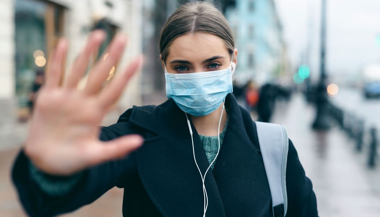 Frau mit Maske, hält eine Hand in die Kamera.