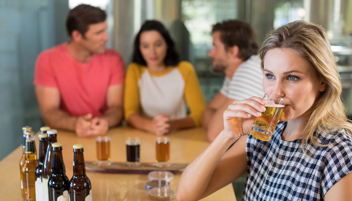 Junge Frau trinkt ein Glas eines alkoholischen Getränks und schaut dabei nachdenklich in die Ferne. Ihre Freunde im Hintergrund unterhalten sich. 