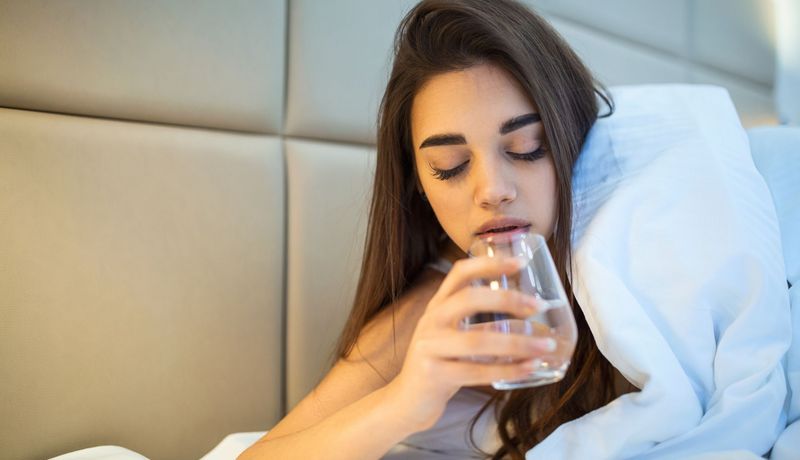 Junge Frau nimmt im Bett eine Schlaftablette mit einem Glas Wasser.