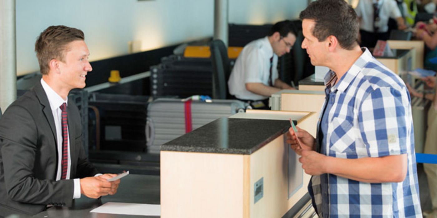 Mann ca. Mitte/Ende 30, in blauweiß kariertem Freizeithemd am Check-in-Schalter im Flughafen