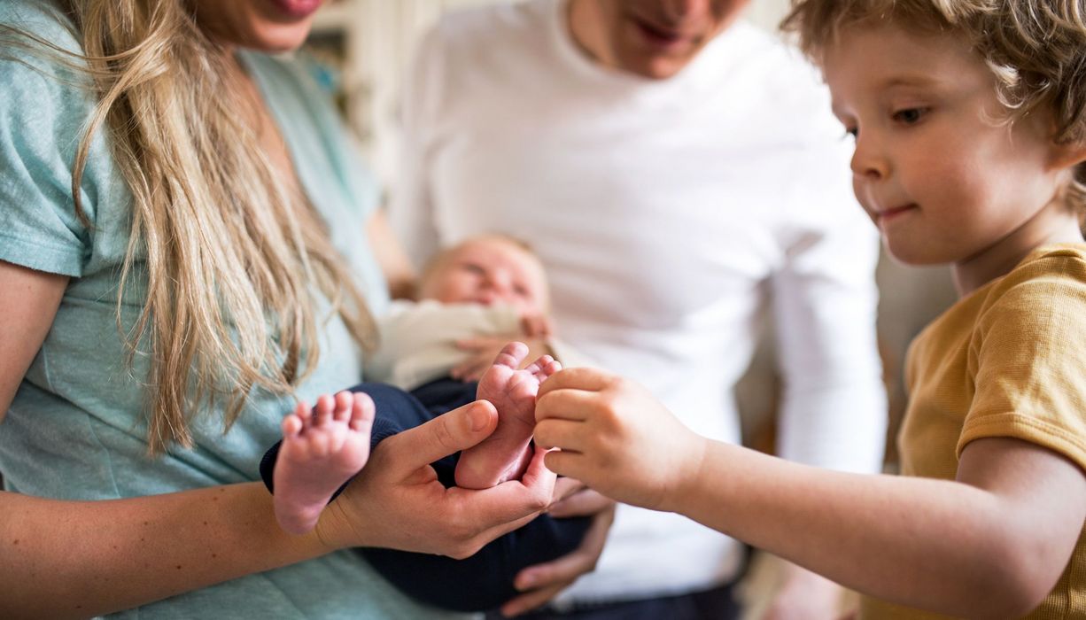 Junge Familie, Mutter hält Neugeborenes im Arm, kleiner Bruder staunt.