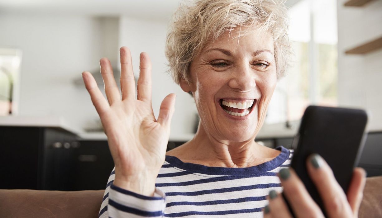 Ältere Frau, lacht in ihr Smartphone und winkt.