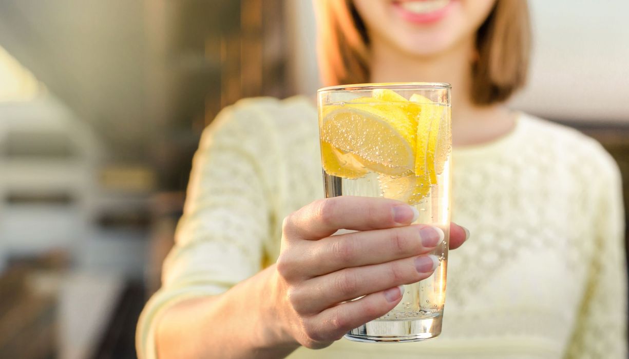 Frau, hält ein Glas mit Wasser und Zitronen in die Kamera.