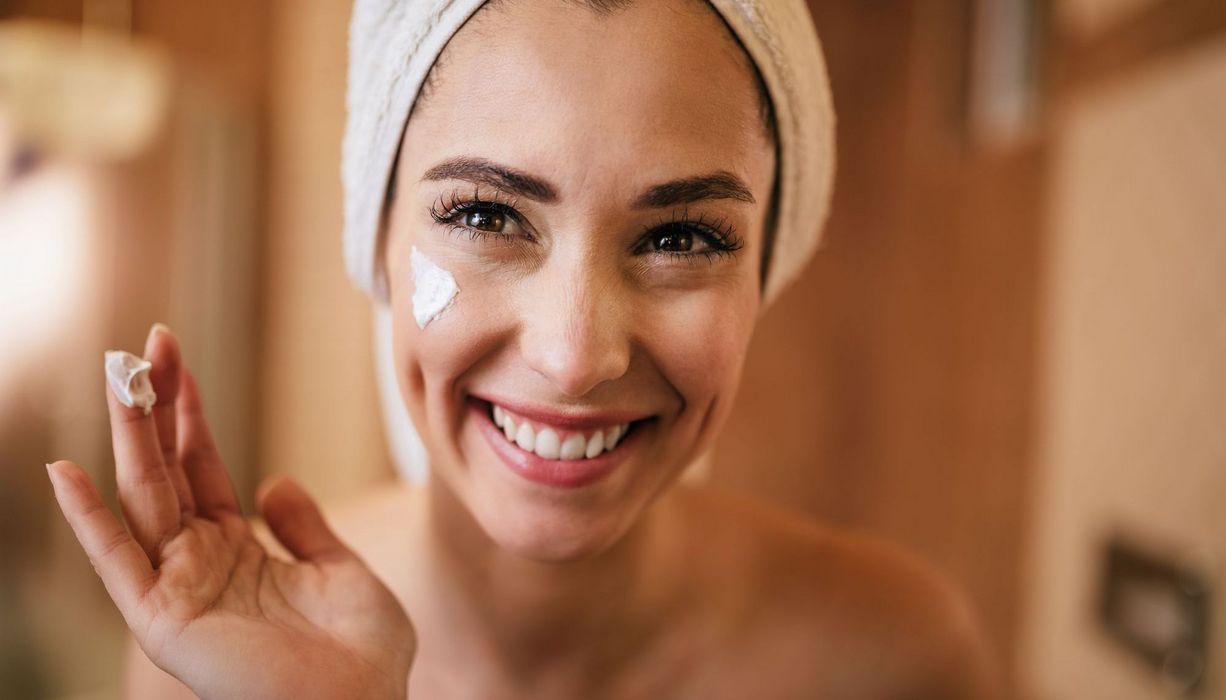 Junge Frau, Haare zu einem Handtuch-Turban gebunden, cremt sich das Gesicht ein.